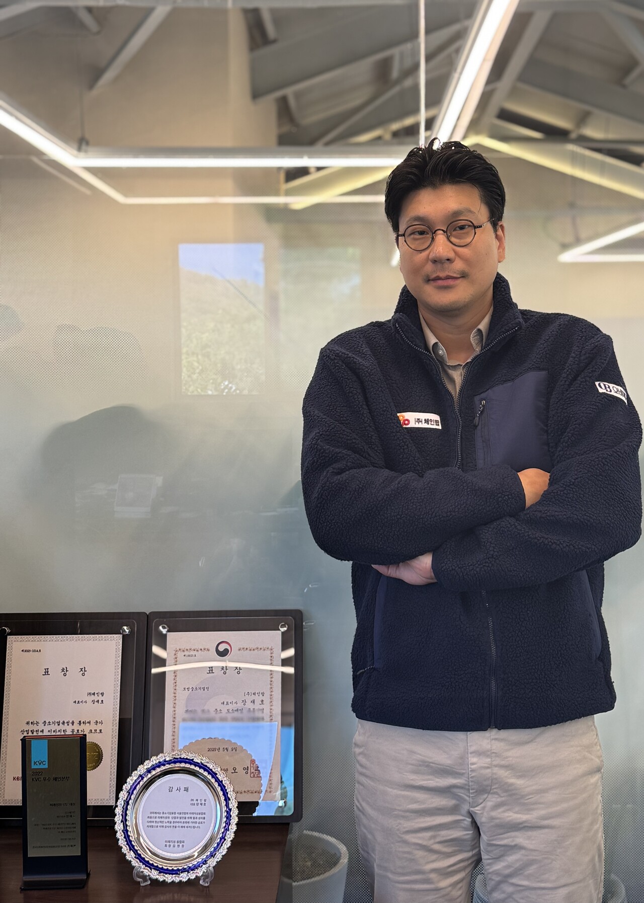 장재호 대표가 체인팝 사무실에서 포상장과 상패 앞에 서 있다. 체인팝은 중소벤처기업부 및 중소기업중앙회로부터 산업 발전 공로를 인정받아 다수의 표창을 수상했다.